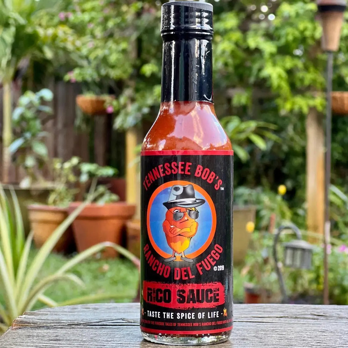 Bottle of Tennessee Bob's Rico Sauce on a wooden surface with a garden background