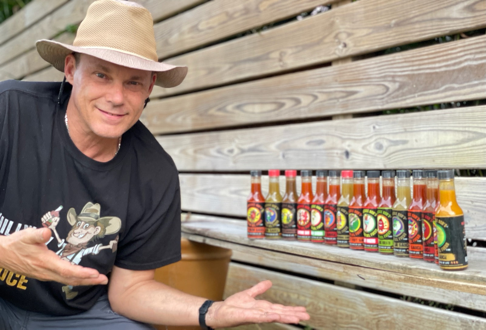 Robert Vasvery sitting on a wooden bench with a row of hot sauce bottles in front of him