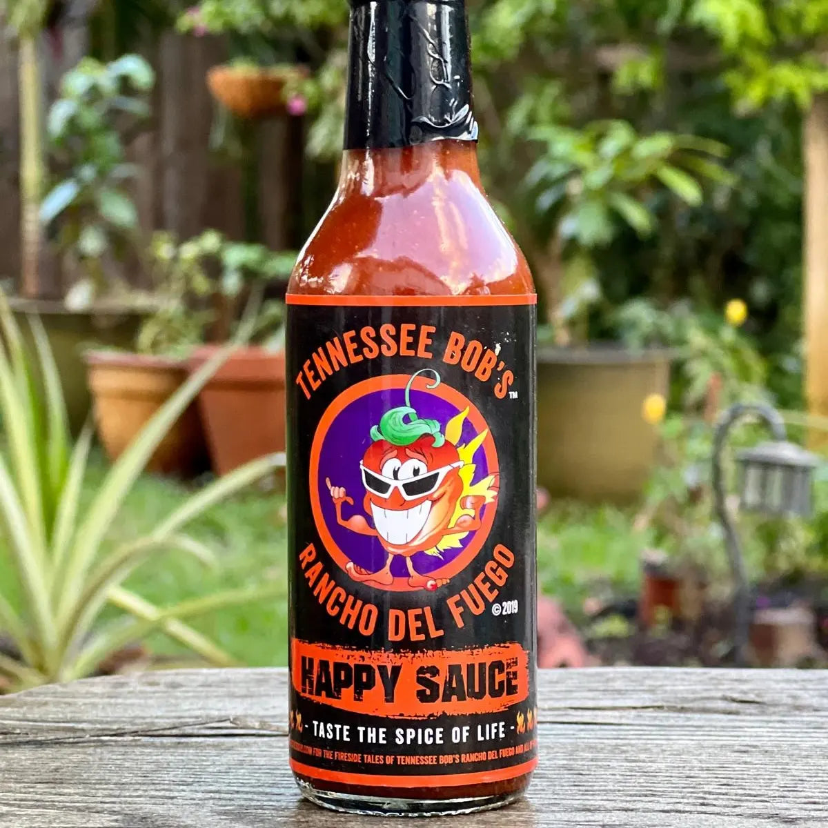 Bottle of Tennessee Bob's Rancho Del Fuego Happy Sauce on a wooden surface with a garden background