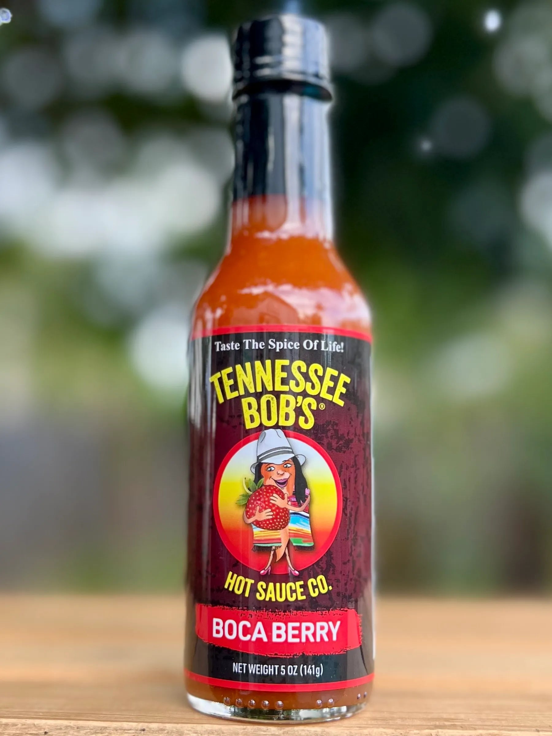 Bottle of Tennessee Bob's Hot Sauce Co. Boca Berry hot sauce on a wooden surface with a blurred green background.