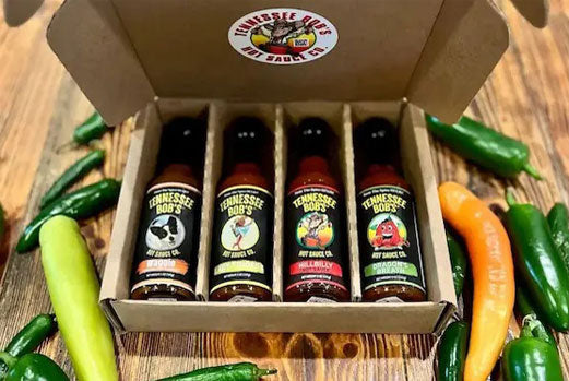 Box of four Tennessee Bob’s hot sauces surrounded by assorted peppers on wooden table.