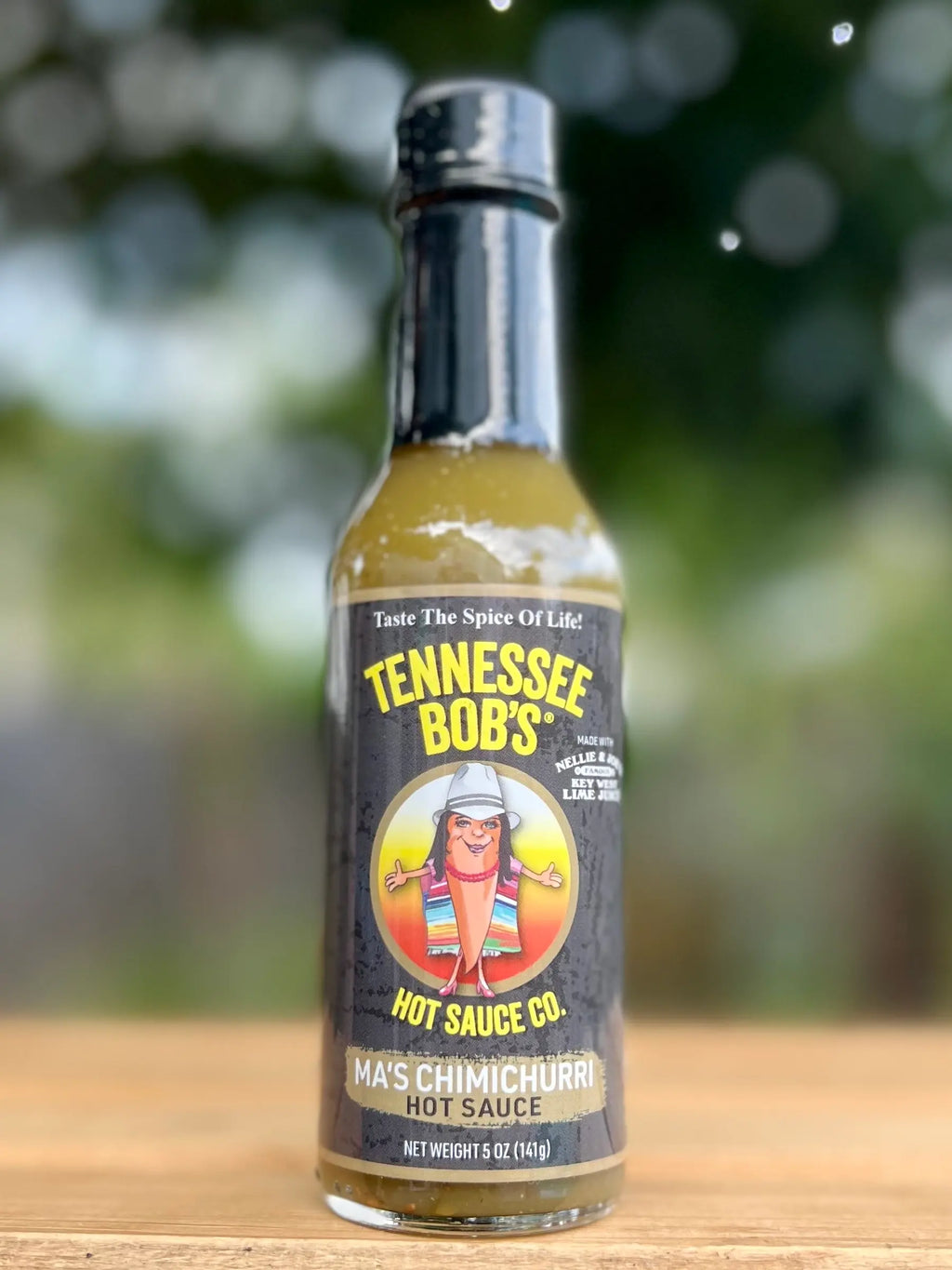 Bottle of Tennessee Bob's Ma's Chimichurri Hot Sauce on a wooden surface with a blurred green background
