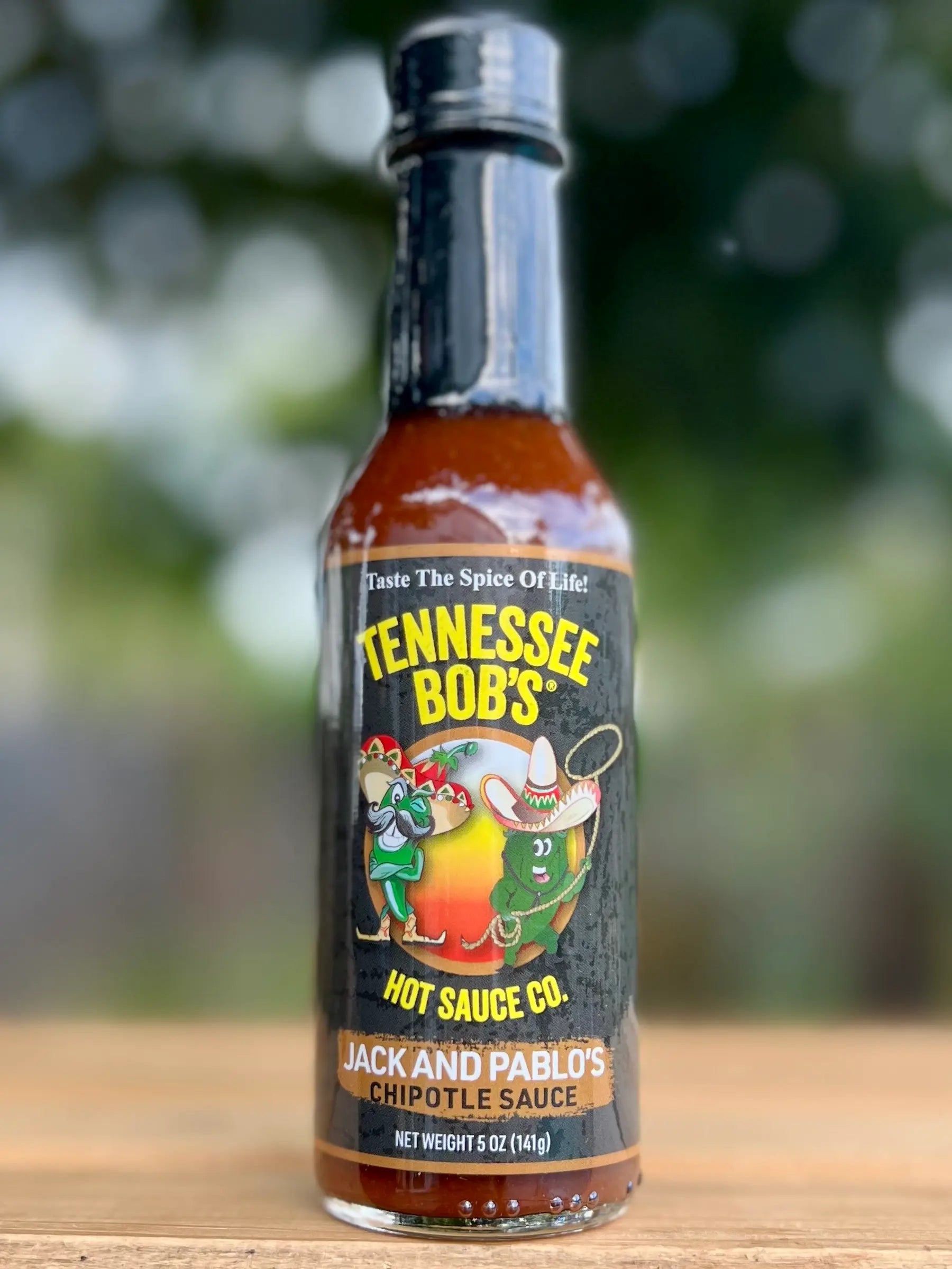 Bottle of Tennessee Bob's Hot Sauce Co. Jack and Pablo's Chipotle Sauce on a wooden surface with a blurred green background.