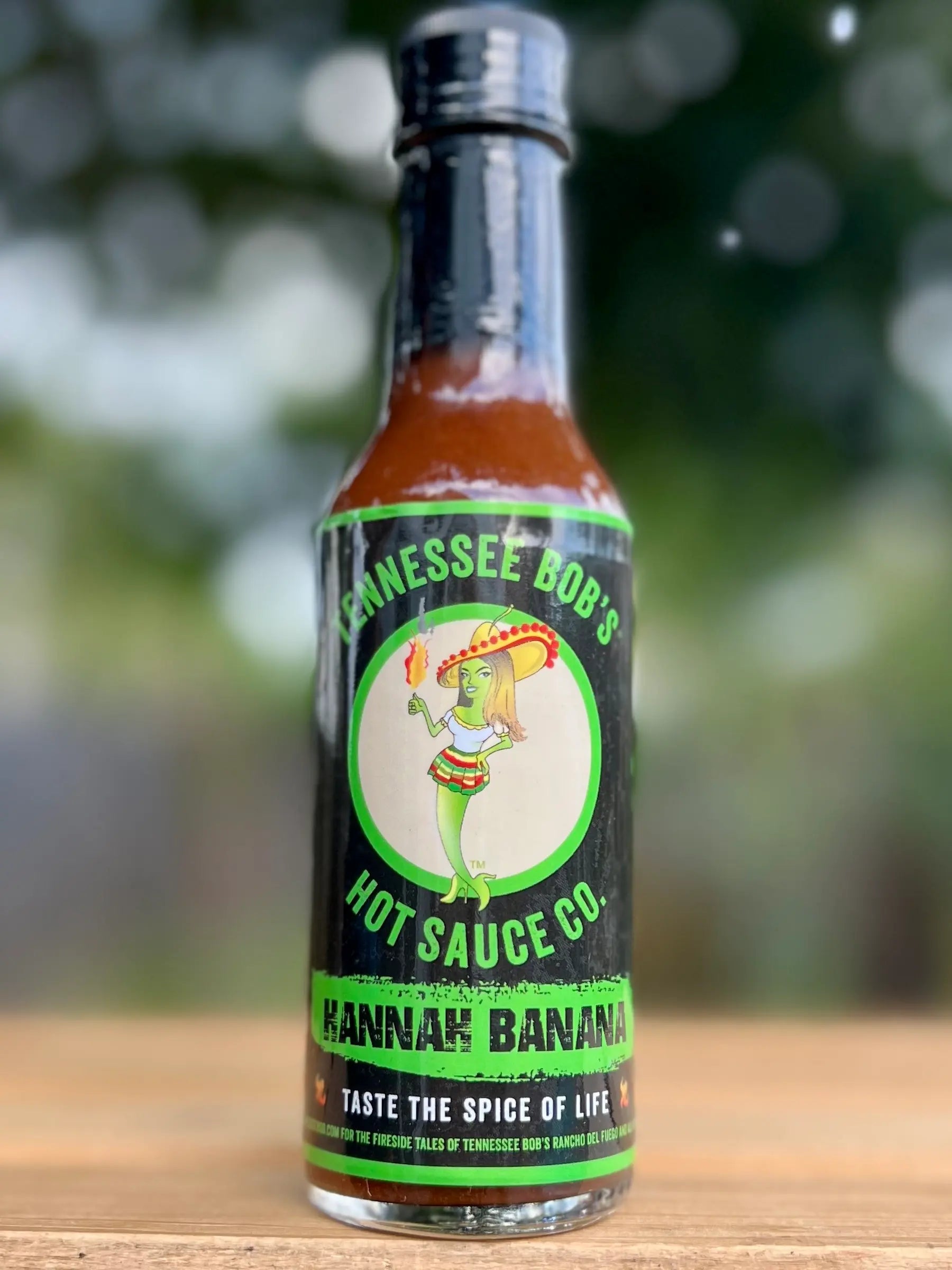 Bottle of Tennessee Bob's Hot Sauce Co. Hannah Banana hot sauce on a wooden surface with a blurred green background.