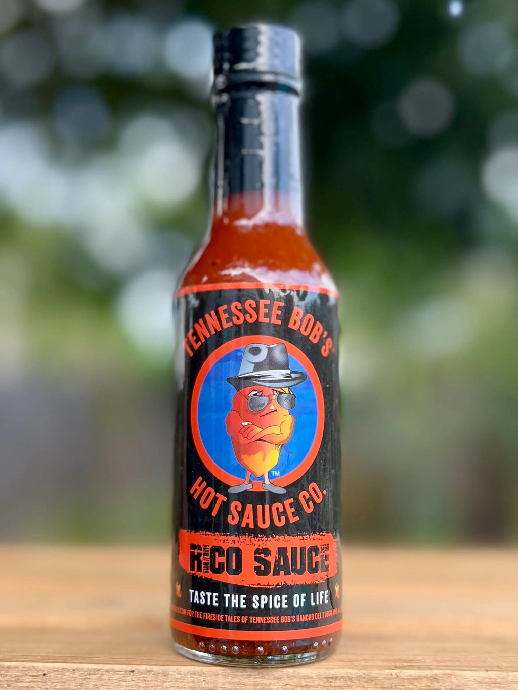 Hot sauce bottle labeled "Tennessee Bob's Hot Sauce Co. Rico Sauce" on a wooden surface with blurred outdoor background.