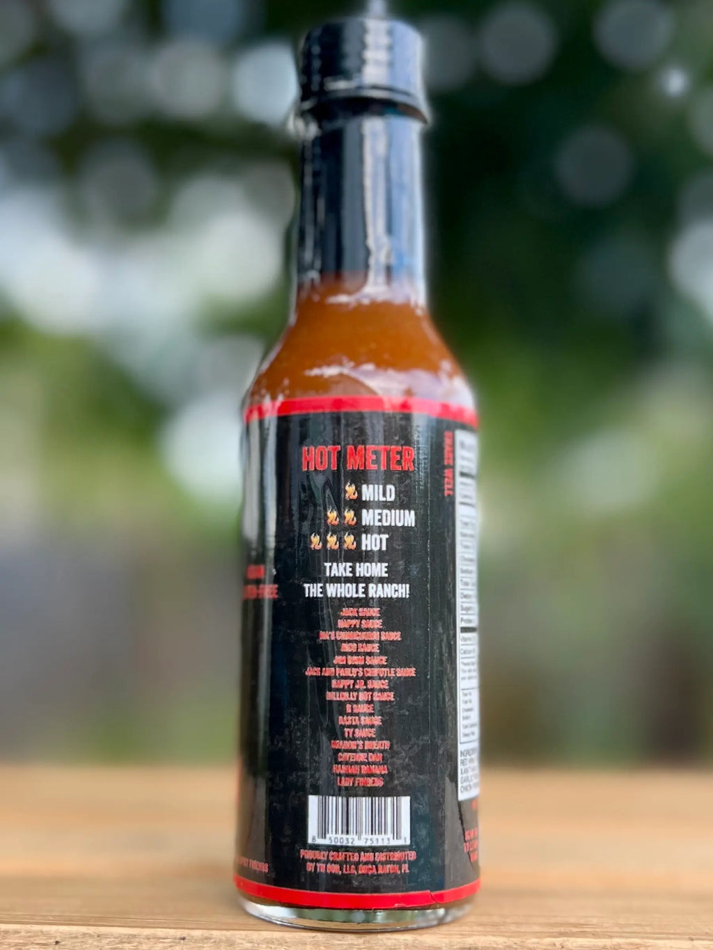 Hot sauce bottle labeled with a "Hot Meter," set on a wooden surface with a blurred outdoor background.