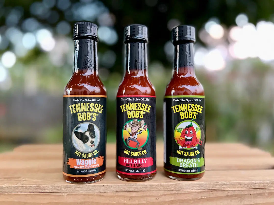 Three bottles of Tennessee Bob's hot sauce on a wooden surface with a blurred outdoor background.