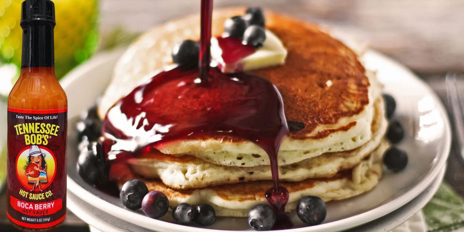 Stack of pancakes with berry syrup and butter, garnished with blueberries, alongside Tennessee Bob's Boca Berry hot sauce bottle.