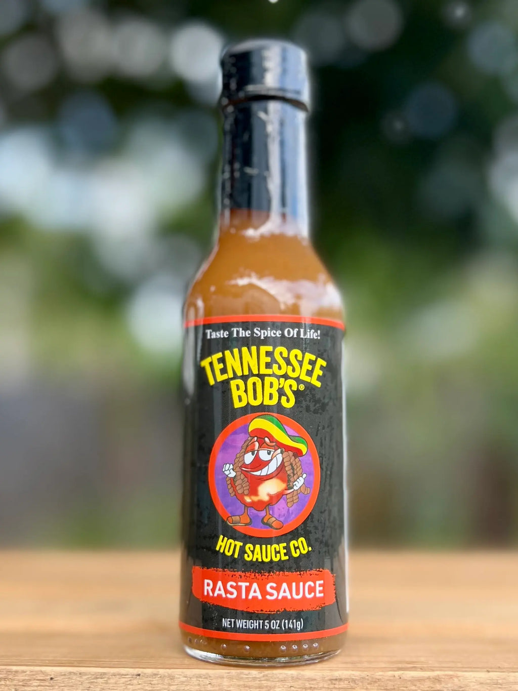 Tennessee Bob's Rasta Sauce bottle on wooden table with blurred outdoor background.