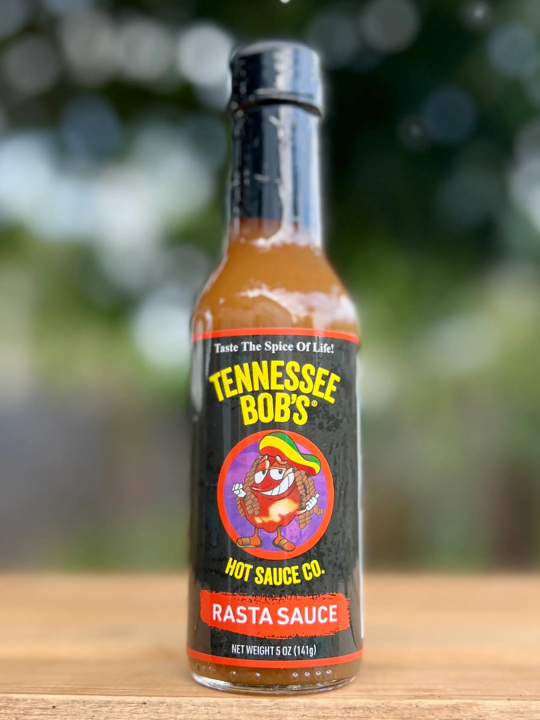 Tennessee Bob's Rasta Sauce bottle on wooden table with blurred outdoor background.